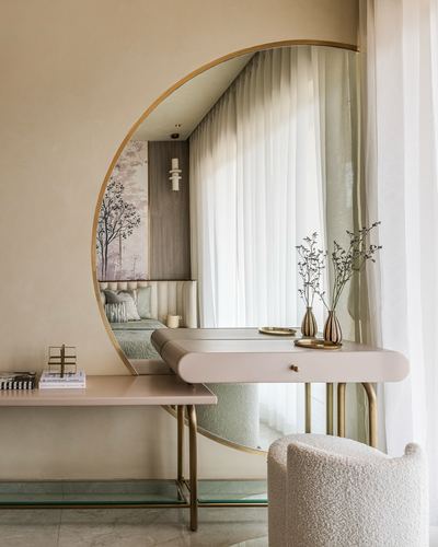 Circular brass-framed mirror above a blush dressing table with bud vases and a boucle armchair