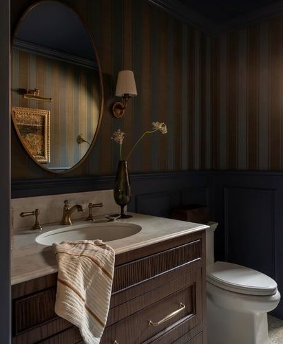 Dark powder room with striped wallpaper, oval mirror, brass sconce, marble-top wood vanity and towel