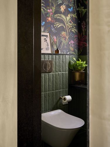 Doorway view into a powder room with green tile, dark floral wallpaper, and a wall-hung toilet