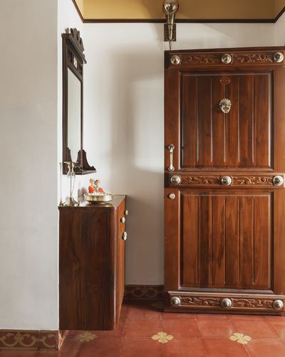 Carved teak entrance door with brass fittings beside a wall-mounted wooden console on red Athangudi tile floor