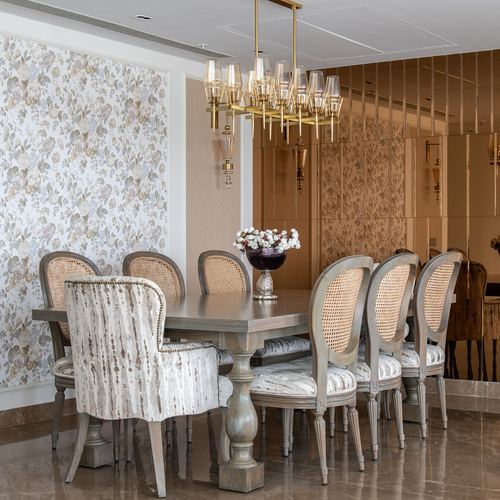 Dining table with cane-back chairs beneath a brass chandelier, floral wallpaper and mirrored panels