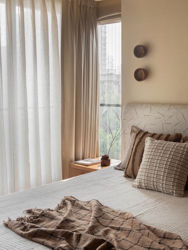 Bedroom corner with sheer curtains, upholstered headboard, and layered textured pillows in warm light