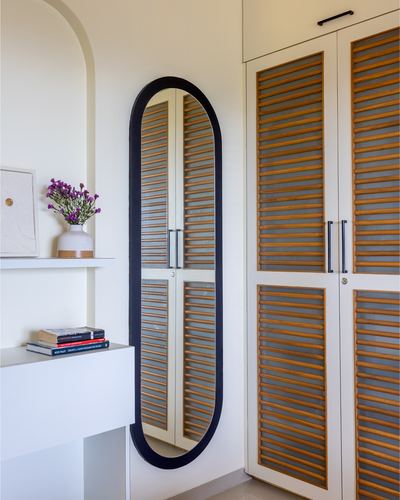 Oval black-framed mirror beside louvered wood wardrobe doors in a white foyer with a vase of purple flowers