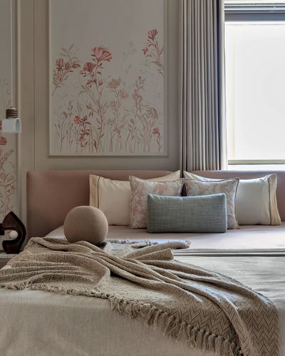 Bed with layered pink and neutral cushions beneath a framed floral artwork and curtained window