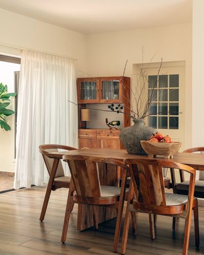 Wooden dining table with carved chairs, glass-front cabinet, and textured vase with dried branches near curtained window