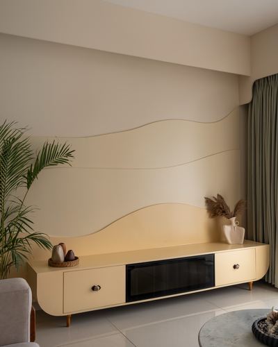 Curved cream wave-patterned wall panelling above a matching rounded tv console with sage curtain and palm plant