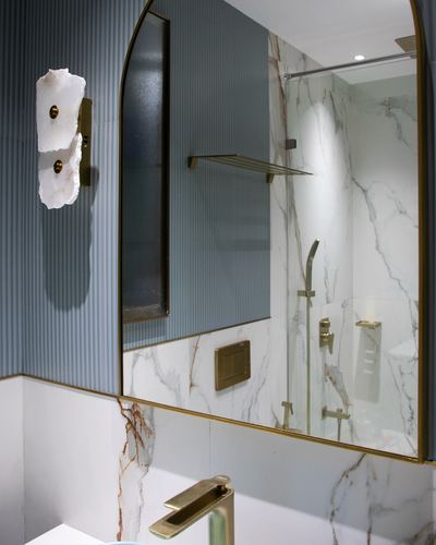 Arched brass mirror reflecting marble shower, alabaster sconce on fluted blue wall