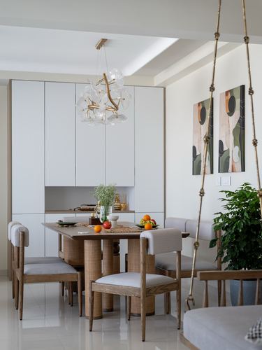 White dining room with wooden cane-base table, sculptural chandelier, and swing chair beside plant