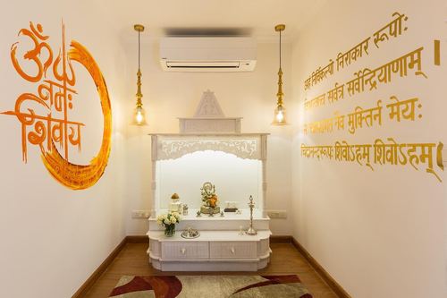 White carved pooja mandir flanked by hanging pendant lights, walls painted with Sanskrit calligraphy