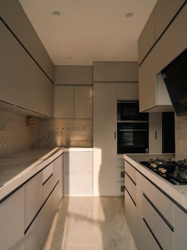 Galley kitchen with taupe cabinetry, stone counters, patterned tile backsplash, and afternoon light on the floor