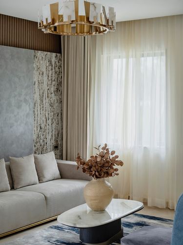 Living room corner with beige velvet sofa, marble coffee table, and brass chandelier by sheer-curtained window