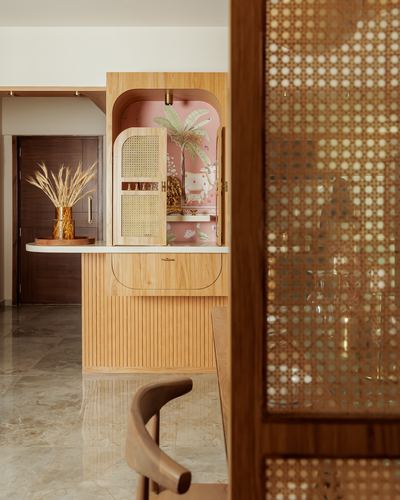 Wooden pooja unit with arched pink-lined niche and cane door, seen through a caned screen partition.