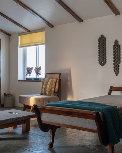 Sunlit studio bedroom with exposed timber beams, cane chair, carved wall panels, and teal-draped bed