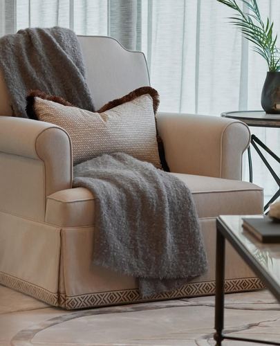 Beige skirted armchair with grey throw and fringed cushion beside a marble-top side table