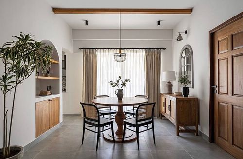 Round wood dining table with cane chairs beneath a wire pendant light, arched niche shelving beside it