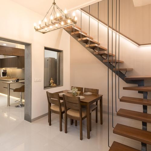 Dining table under a floating wood-and-steel staircase beside a kitchen and lit wall niche