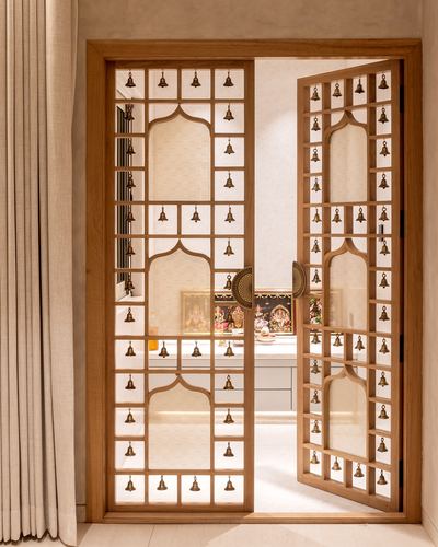 Wooden jali doors with hanging brass bells open to reveal a pooja room altar with deity idols