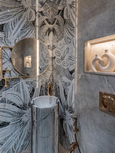 Powder room with mirrored mosaic wall, round brass mirror, fluted stone pedestal basin, and lit stone niche
