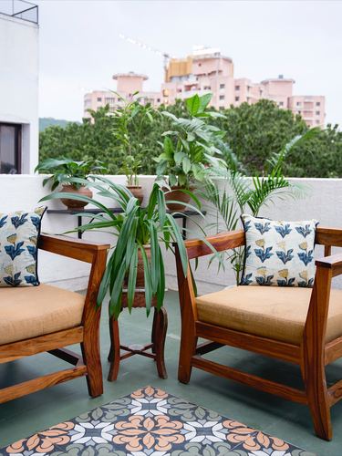 Two wooden armchairs with printed cushions on a balcony surrounded by potted plants, city skyline behind