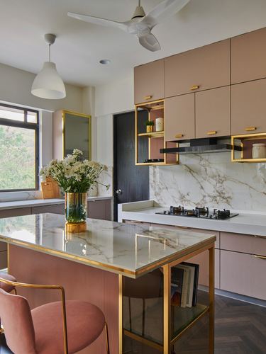 Pink and gold kitchen with marble island, open brass shelving, and herringbone wood floor