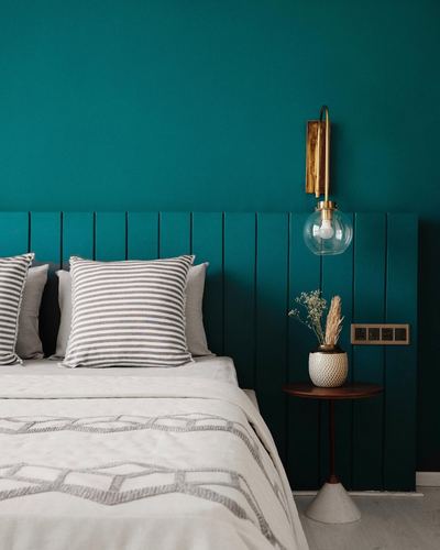 Teal bedroom with wood panel headboard, brass sconce, and round side table with vase.