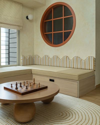 Corner banquette with striped scalloped cushions below a round terracotta window, round wood table with chess set in front.