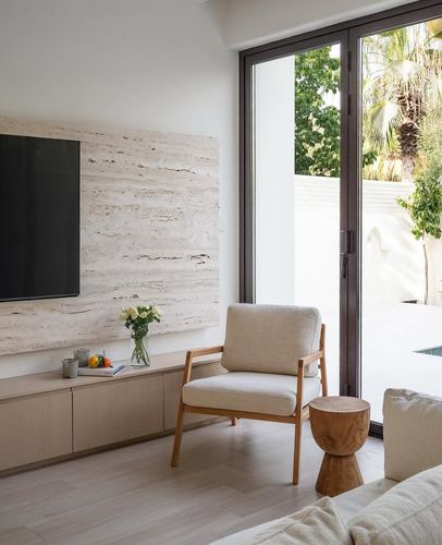 Minimalist living room with travertine tv wall, cane-and-linen armchair, and glass doors opening to a garden