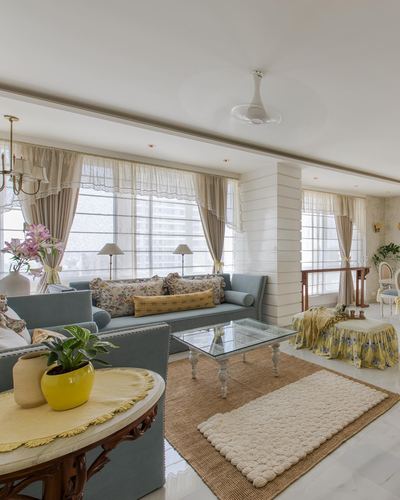 French-style living room with blue sofas, floral cushions, layered rugs, and sheer curtained windows