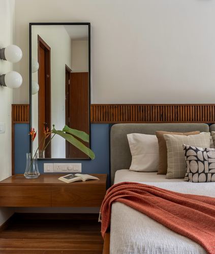 Bedroom corner with teak-framed mirror, globe sconces, floating nightstand, and bed with terracotta throw