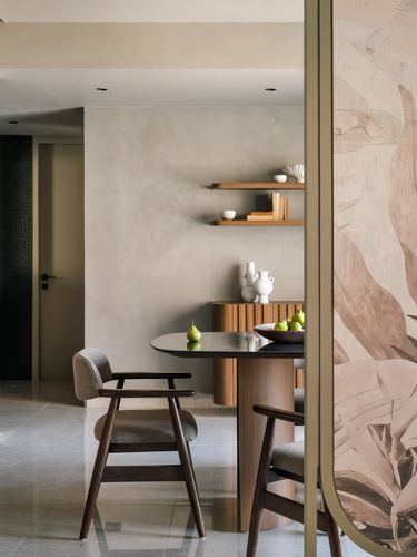 Round wooden dining table with cane chairs beside floating shelves and a floral screen