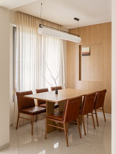 Wooden dining table with tan leather chairs beneath a linear pendant light against sheer curtains