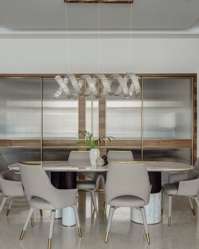 Oval stone dining table with beige chairs under a ribboned pendant light, ribbed glass cabinetry behind