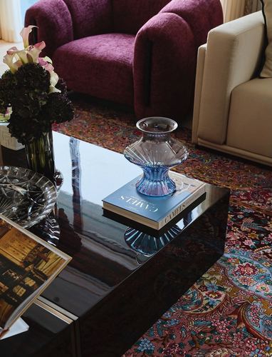Glass vase and book on glossy black coffee table beside burgundy armchair and patterned rug