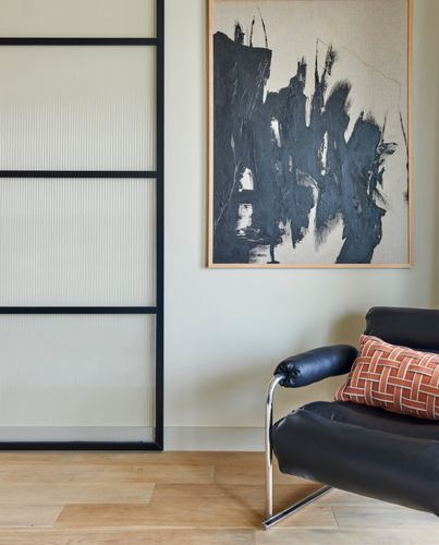 Black leather chair with terracotta pillow beneath large abstract painting, reeded glass door beside.