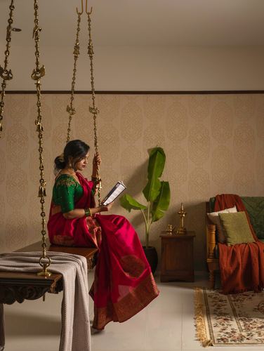 Woman in red saree reading on brass jhula in warmly lit South Indian living room
