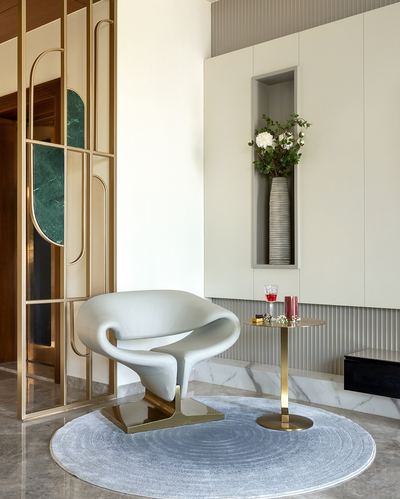 Cream sculptural armchair with brass base beside a round brass side table and niche wall in a marble-floored foyer