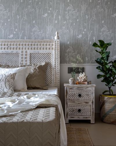 Carved whitewashed wood headboard and matching nightstand beside a fiddle leaf fig plant in a floral-papered bedroom.