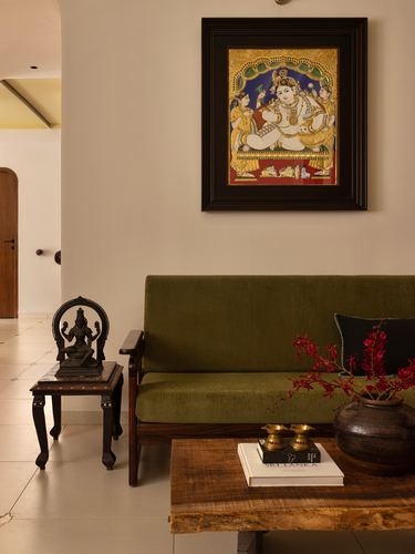 Olive velvet wooden sofa beneath a framed Tanjore painting, with brass deity figurine on side table