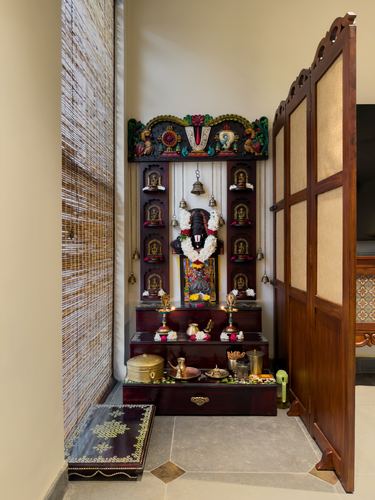 Traditional carved wooden pooja mandir with bells, garland, and brass diyas beside a woven screen door