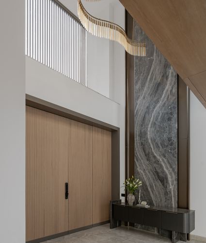Double-height foyer with ribbed wood doors, veined stone wall, and cascading gold chandelier