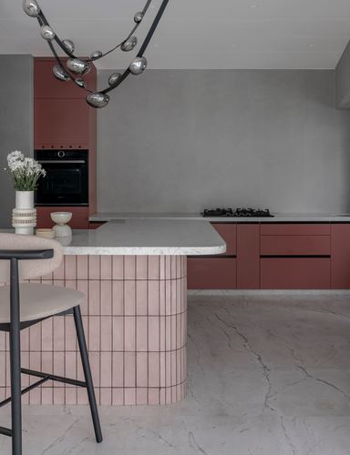 Curved blush-tiled kitchen island beside terracotta cabinetry and grey stone floor under a beaded pendant light
