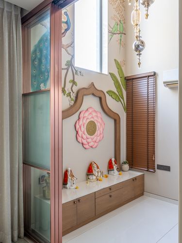 Pooja unit with carved arched frame, pink lotus wall art, deity idols, and peacock mural beside sliding glass door