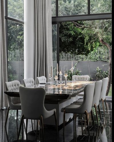 Dining table with tufted chairs and gold legs facing floor-to-ceiling glass wall overlooking garden