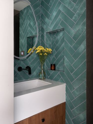 Bathroom vanity with teal herringbone tile wall, round mirror, and yellow flowers in glass vase