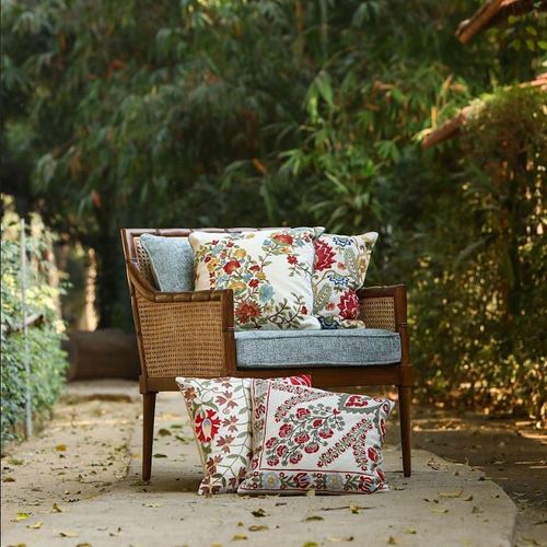 Cane-backed wooden armchair with floral embroidered cushions on a stone path amid garden greenery.