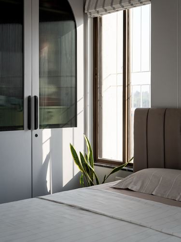 Sunlit bedroom corner with fluted wardrobe, window, plant and beige upholstered headboard