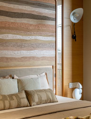 Bed with layered cushions against a wave-patterned stone headboard wall and wood-clad wall with sconce