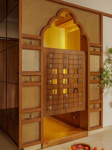 Wooden pooja unit with scalloped arch, grid door panel, brass bells, and warm interior glow