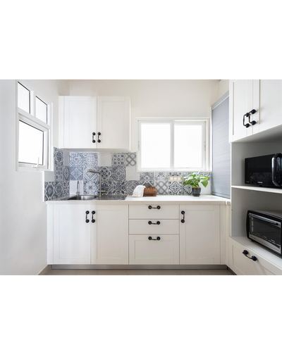White L-shaped kitchen with patterned blue-grey tile backsplash and black hardware, lit by two windows