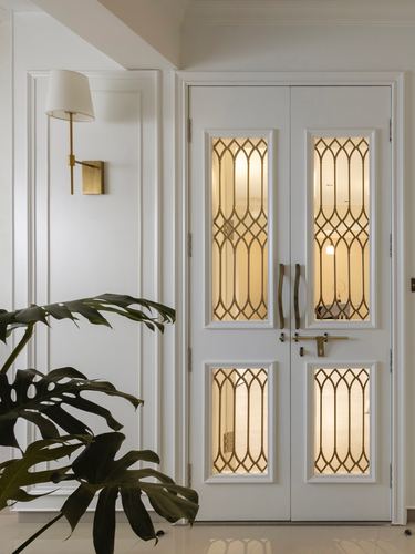 White double door with brass lattice glass panels, brass sconce, and monstera plant in foyer
